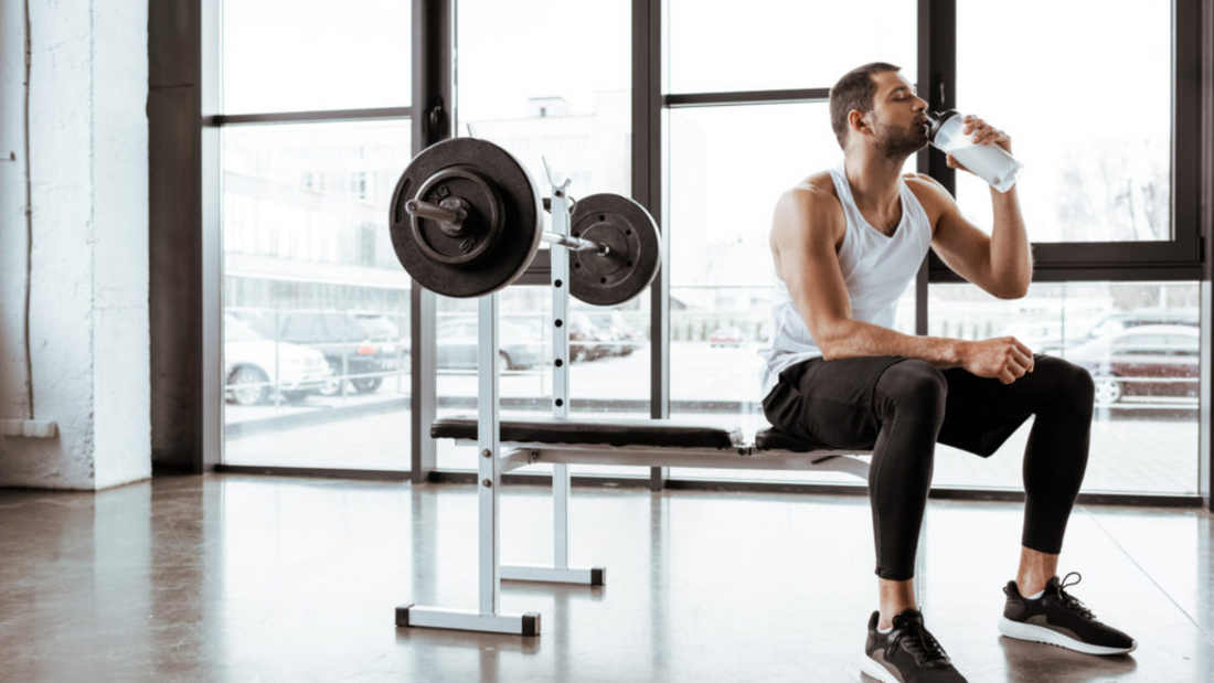 Man sitting on a bench in a gym, drinking water next to a barbell, focusing on fitness and hydration.